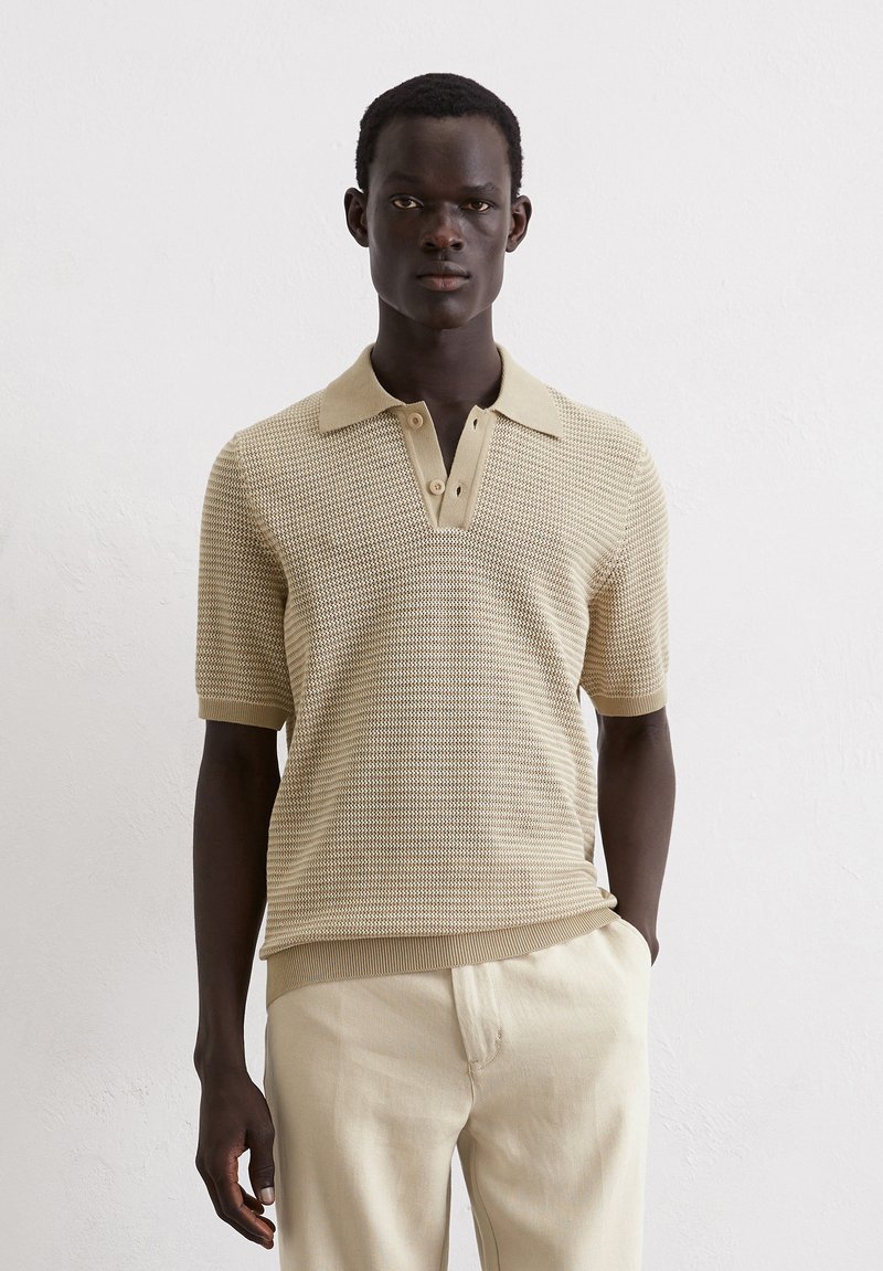 Beige knitted polo shirt with a textured pattern, buttoned collar, and short sleeves, paired with light beige trousers.