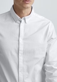 Chemise blanche à boutons avec un petit col, fabriquée en coton. Elle présente une texture lisse et une fermeture à boutons classique sur le devant.