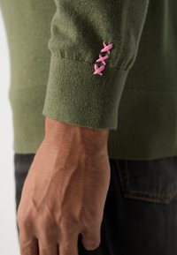 Pulover verde din tricot cu manșete cu danteluri, având trei accente brodate în formă de X roz. Prim-plan al detaliului brațului și al mâinii, material texturat.