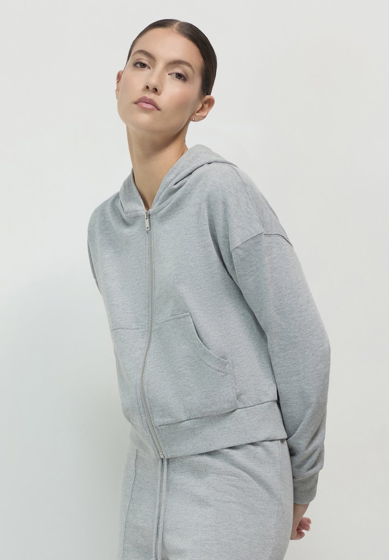 Sweat à zip gris en tissu doux, featuring une capuche, des poches latérales et des poignets côtelés. Le design est décontracté et casual.