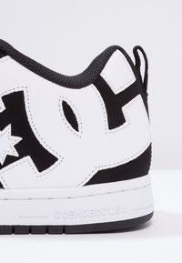 Valkoinen lenkkarit mustilla yksityiskohdilla, joissa on pehmustettu kaulus, kuvioitu kumipohja, suuri brändäys ja vastaväriset materiaaliosiot.