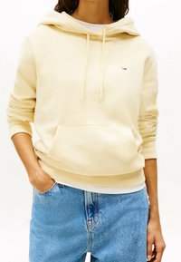 Keltainen hupullinen collegepaita, jossa on etutasku, ribbaus hihansuissa ja helmassa, yhdistettynä vaaleansinisiin farkkuihin. Rinnassa on pieni logo.