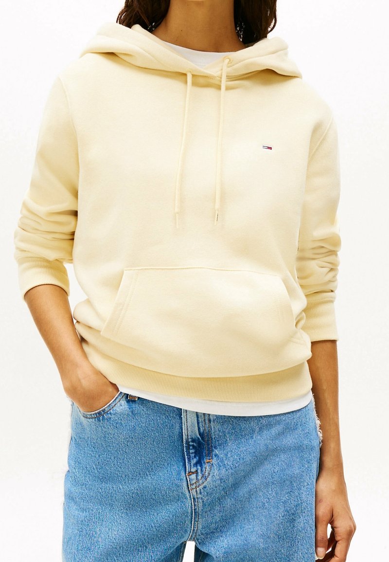 Keltainen hupullinen collegepaita, jossa on etutasku, ribbaus hihansuissa ja helmassa, yhdistettynä vaaleansinisiin farkkuihin. Rinnassa on pieni logo.