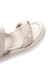 Beige sandaal met een gevlochten band, soepel leren bovenwerk, gevoerd voetbed en een witte rubberen zool. Bevat minimale stikdetails.