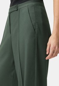 Pantalones verdes de pierna ancha hechos de tela suave, con cierre de botones, frente plisado y bolsillos laterales. Detalles texturizados visibles.
