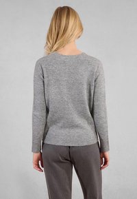 Persona bionda in piedi di fronte a un muro grigio, che indossa un maglione di maglia grigia e pantaloni grigi scuri, con le mani rilassate ai fianchi.