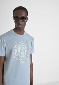 Camiseta de algodón azul claro con un diseño recortado de la cabeza de un león en el frente. La camiseta tiene un cuello redondo y mangas cortas.