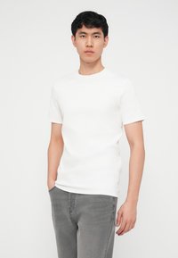 Νέος άνδρας με κοντά σγουρά μαλλιά, φορώντας απλό λευκό κοντομάνικο πουκάμισο και γκρι τζιν, στέκεται μπροστά σε ένα απλό λευκό φόντο.