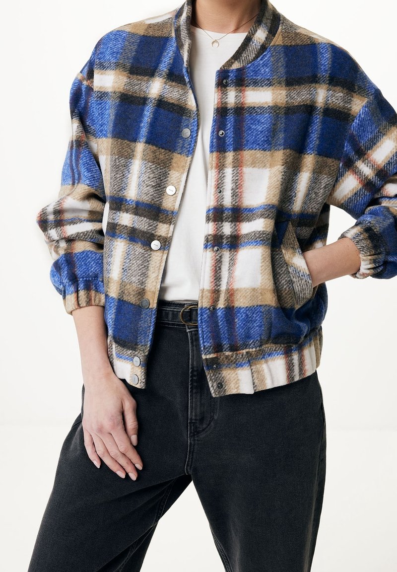 Personne portant une veste à carreaux bleus, bruns et blancs sur une chemise blanche avec un jean noir, une main dans la poche et l'autre posée sur la jambe.