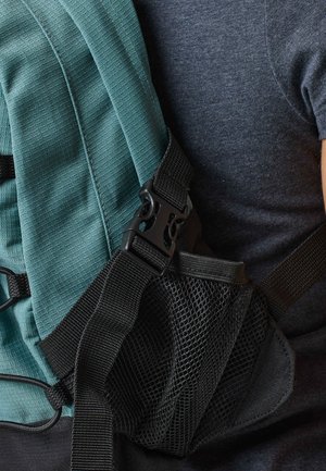 Close-up van een schouderriem van een blauwe rugzak met een zwarte gesp en een gaasvak, gedragen over een donkergrijs shirt.