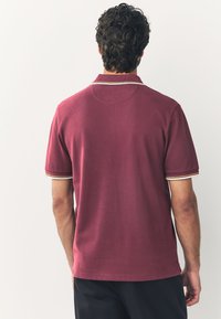Bordeauxrotes Poloshirt mit kurzen Ärmeln, mit geripptem Kragen und mehrfarbigen Streifenakzenten an den Ärmelbündchen. Glatte Textur, klassischer Schnitt.
