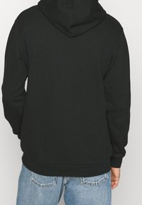 Černá mikina s kapucí vyrobená z měkké látky, volného střihu, s nastavitelnou kapucí na šňůrku a žebrovanými manžetami a lemem. Nenošena s světlemodrými džíny.