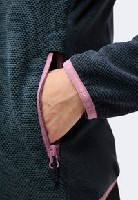 Main insérée dans la poche zippée latérale d'une veste texturée foncée avec fermeture éclair et bordure de manchette roses.