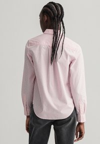 Een persoon met gevlochten haar draagt een roze geruit overhemd en grijze broek, met de rug naar de camera gekeerd tegen een minimalistische grijze achtergrond.