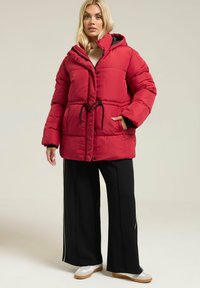 Veste rembourrée rouge avec capuche, taille ajustable par cordon, boutons pression et poches latérales. Portée par-dessus un pantalon large noir et des baskets blanches.