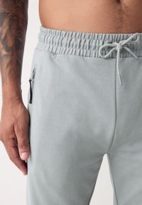 Pantaloni de trening gri deschis cu talie elastică, șnur și buzunar lateral cu fermuar. Textură netedă cu un design cu croială strâmtă.