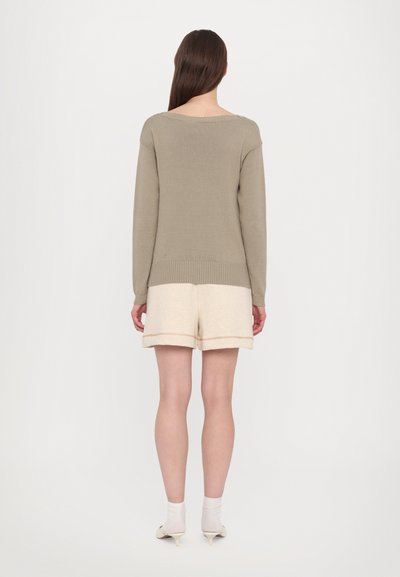 Ανοιχτός μπεζ πλεκτός πουλόβερ με φαρδιά λαιμόκοψη, σε συνδυασμό με κρεμ σορτσάκι με διπλωμένο στρίφωμα και διακριτική υφή.
