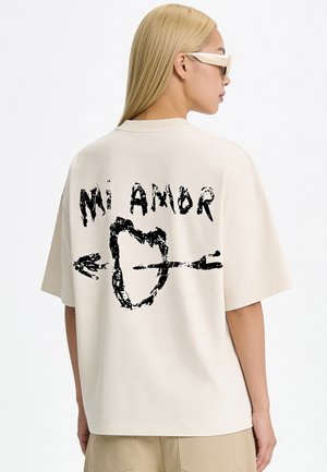 Blond persoon met beige zonnebril en los beige T-shirt met een zwarte afbeelding van een hart doorboord met een pijl en tekst "Mi Amor" op de achterkant.
