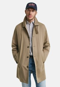 Beige trenchcoat met een kraag, voorknopsluiting en zijzakken, gelaagd over een blauw-wit gestreept shirt, gecombineerd met denim jeans.