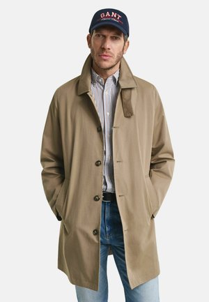 Beige Trenchcoat mit Kragen, Vorderknöpfen und seitlichen Taschen, über einem blau-weißen gestreiften Hemd getragen, kombiniert mit Jeans.