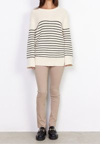 Cremfarbener Pullover mit schwarzen Streifen, lockere Passform und weiten Ärmeln, kombiniert mit beigen, schmalen Hosen und schwarzen Slippern mit Hardware-Akzent.