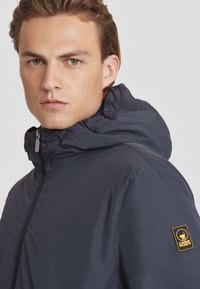 Marineblauwe pufferjack met hoge kraag, ritssluiting aan de voorkant en een logo-opdruk op de mouw; gladde textuur, lichtgewicht materiaal.