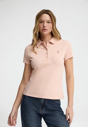 Kvinde iført lys lyserød kortærmet polo t-shirt med knapstolpe og mørkeblå jeans, stående mod en ensfarvet lys baggrund.