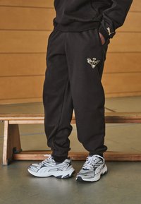Black sweatpants with elastic cuffs, featuring a gold logo on the left thigh, paired with white and grey sneakers with black accents.