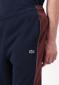 Mørkeblå og bordeaux sweatpants med elastisk talje, der har et lille grønt Lacoste-logo på venstre lår og kontrasterende sidelister.