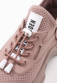 Chaussure rose avec une tige en maille, des renforts en caoutchouc, des lacets élastiques aux embouts blancs et une étiquette avec un texte noir sur fond blanc.