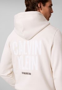 Homme portant un sweat à capuche écru, montrant son dos avec un grand "Calvin Klein" estompé et un texte plus petit "Established 1968" en dessous.