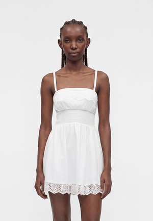 Frau mit geflochtenem Haar, die ein weißes ärmelloses Kleid mit Spitzenbesatz, geraffter Taille und quadratischem Ausschnitt trägt, steht vor einem schlichten Hintergrund.