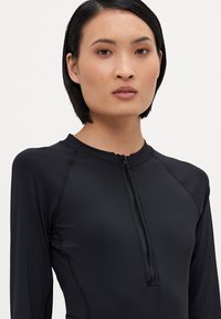 Camisa de proteção de manga longa preta com colarinho alto, apresentando um fecho frontal e costura em contraste. Feita de um tecido suave e elástico.