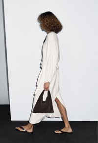 Donna con capelli ricci che cammina, indossa un vestito bianco a maniche lunghe con una spacco laterale, porta una borsa nera e sandali bassi neri.