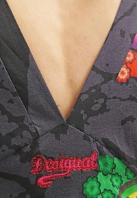 Szary top z dekoltem w serek z kwiatowym wzorem, z czerwonymi, zielonymi i purpurowymi akcentami. Zawiera haftowane logo "Desigual" przy dekolcie.