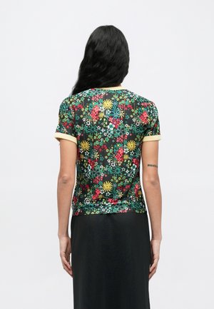 Femme aux longs cheveux noirs portant une chemise à manches courtes avec des motifs floraux colorés et une jupe noire, vue de dos.