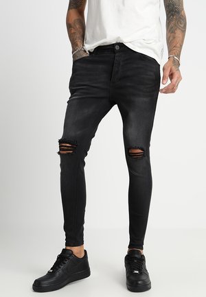 Mand iført sorte, slidte, tætsiddende jeans, sorte sneakers, hvid t-shirt, med tatoverede arme og hænder i lommer, stående på en grå gulv.