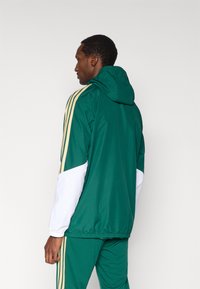 Vihreä hupparillinen takki, jossa valkoisia ja kultaisia yksityiskohtia, sileä pinta ja joustavat hihansuut. Yhdistetty samaa vihreää oleviin housuihin.