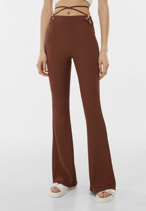 Pantaloni marroni a vita alta e dalla gamba svasata con occhielli laterali e cinturini avvolgenti, indossati con sandali platform bianchi.