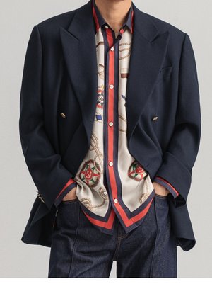 Mann trägt einen marineblauen Blazer über einem cremefarbenen Hemd mit roten und marineblauen Streifen, kombiniert mit dunklen Jeans, Hände in den Taschen.