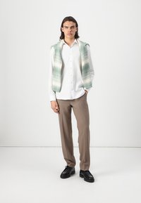 Chemise blanche à boutons, pantalon beige ajusté et un pull à motif vert menthe drapé sur les épaules. Des chaussures noires complètent l'ensemble.