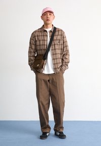 Cămașă maro cu carouri, purtată peste un tricou alb, pantaloni maro și o pălărie roz. Încălțăminte neagră cu dungi albe și un rucsac maro purtat pe umăr.