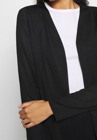 Černý kardigan s dlouhými rukávy a otevřeným předním dílem, ribbované textury, kombinovaný s bílým tričkem pod ním. Hladká látka a uvolněný střih.