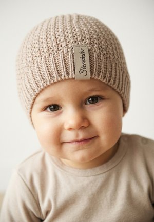 Toddler wearing a beige knitted hat and matching shirt, smiling gently at the camera against a plain light background.