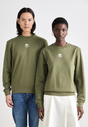 Olijfgroene sweaters met geribbelde manchetten, voorzien van een wit Lacoste-logo. Draag ze met blauwe jeans en een witte rok. Soepele katoenen stof.