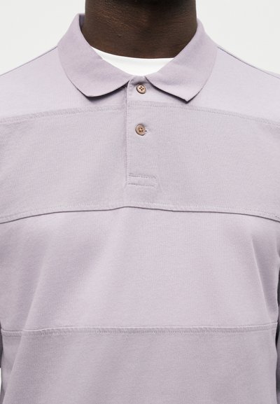 Άνδρας που φοράει ένα ανοιχτό μωβ πόλο με δύο καφέ κουμπιά και ένα λευκό υποκάμισο που φαίνεται στο γιακά.