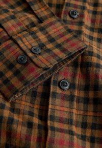 Camicia di flanella marrone con motivo a quadri in arancione, rosso e nero. Presenta bottoni neri sul davanti e sui polsini.