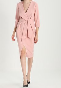 Robe enveloppante rose avec des manches trois-quarts, une taille nouée et une fente jusqu'à la cuisse, fabriquée en tissu lisse. Portée avec des talons hauts noirs.
