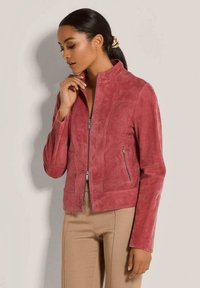 Rosa Wildlederjacke mit Stehkragen, strukturierten Details an den Seiten, Frontreißverschluss und flachen Nähten. Kombiniert mit braunen Hosen.