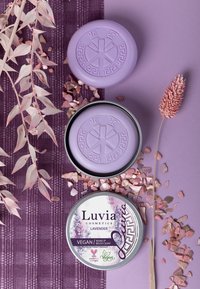 Violetti pyöreä saippua, jossa on kohokuvioitu design ja laventelin tuoksu, tinan vieressä, jossa lukee "Luvia Cosmetics." Ympärillä on vaaleanpunaisia lehtiä ja kiteitä.